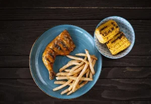 1/4 chicken leg and thigh with fries and grilled corn on a dark plate